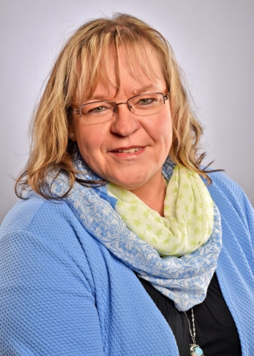 Portrait Corina Fitzner Ein Porträtfoto einer freundlich lächelnden Frau mit längeren, blonden Haaren und einer Brille. Sie trägt eine hellblaue Strickjacke über einem dunklen Oberteil und dazu einen voluminösen, hellgelben Schal. Der Hintergrund ist in einem neutralen, hellen Grau gehalten. Das Bild wirkt herzlich, kompetent und professionell.