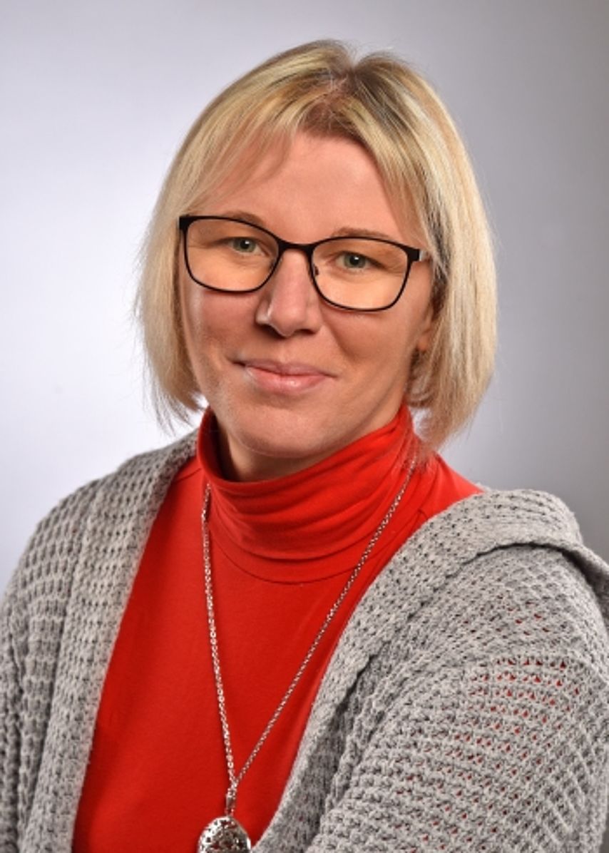 Portrait Jana Bachmann Ein Porträtfoto einer freundlich lächelnden Frau mit kurzen blonden Haaren und einer schwarzen Brille. Sie trägt einen leuchtend roten Rollkragenpullover und darüber eine grau melierte Strickjacke. Der Hintergrund ist in einem neutralen, hellen Grau gehalten. Das Bild wirkt professionell, sympathisch und einladend.