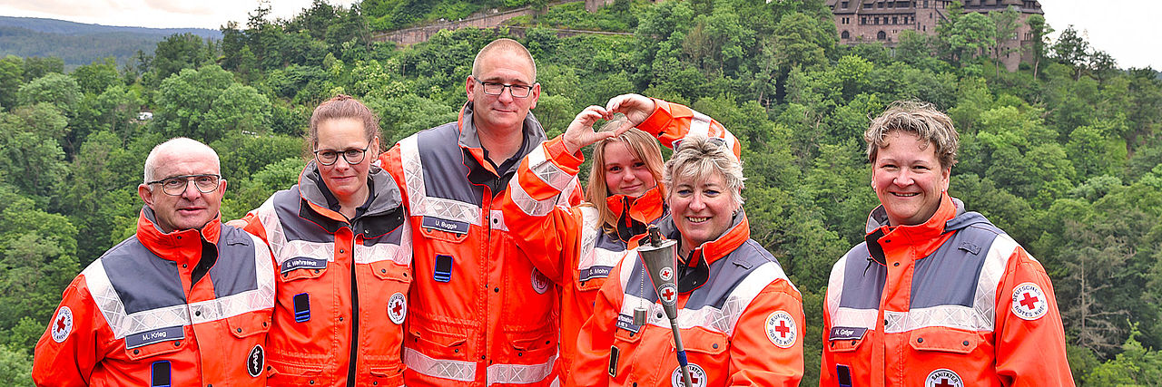 Sechs fröhliche DRK-Einsatzkräfte in orangefarbenen Jacken zeigen Teamgeist und lächeln. Im Hintergrund sind die historische Wartburg und ein bewaldeter Berghang zu sehen.