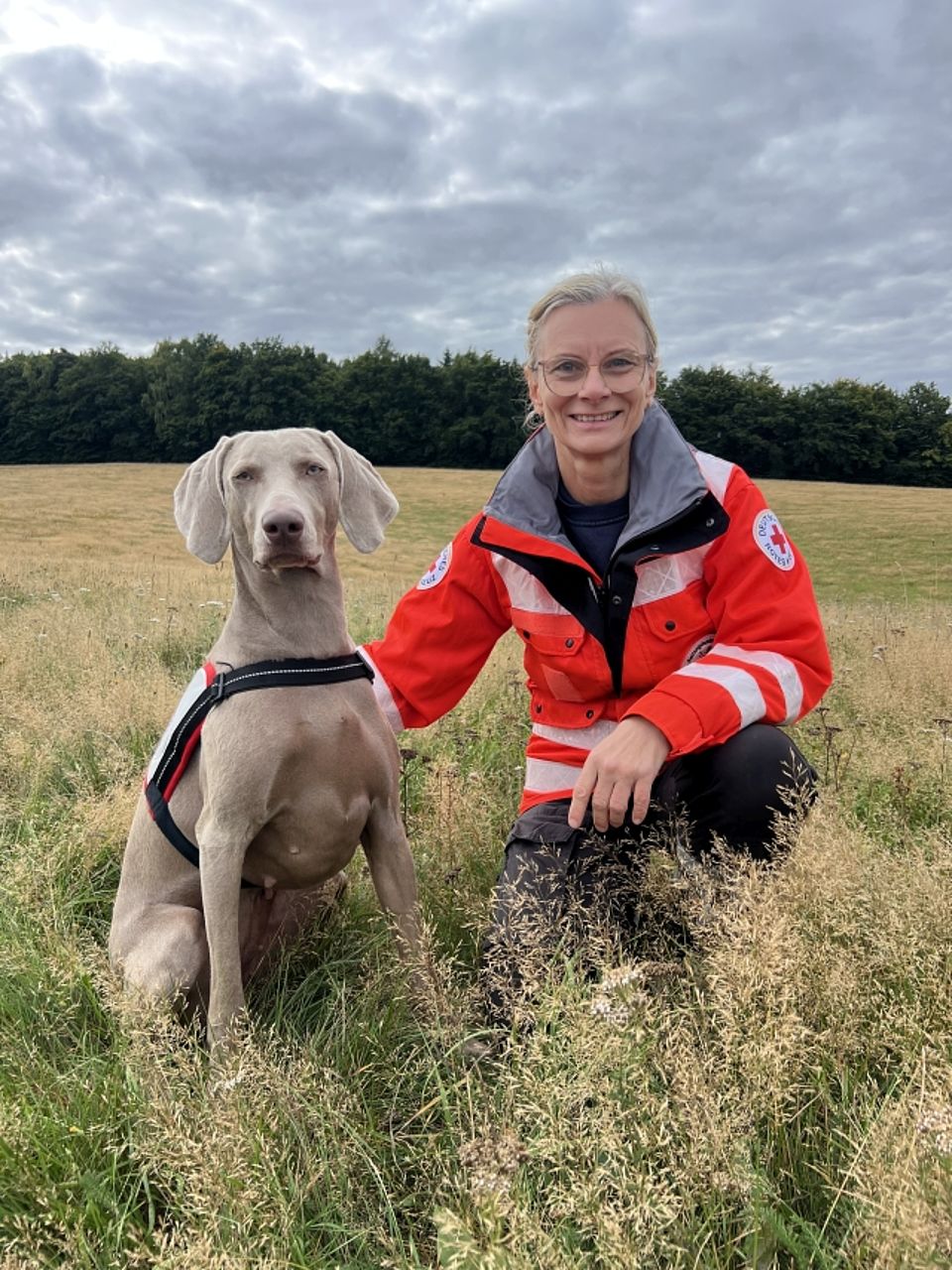 Manuela mit Klara Eine lächelnde, blonde Frau (Rettungskraft) in orange-grauer Einsatzjacke des Deutschen Roten Kreuzes kniet auf einer Wiese neben einem silbrigen Weimaraner Rettungshund. Dieser sitzt ruhig und trägt ein rot-weißes Geschirr mit dem Roten Kreuz. Die Frau legt ihre Hand auf den Rücken des Hundes. Die Szene spielt im Freien vor einem dunklen Waldrand unter einem bewölkten Himmel.