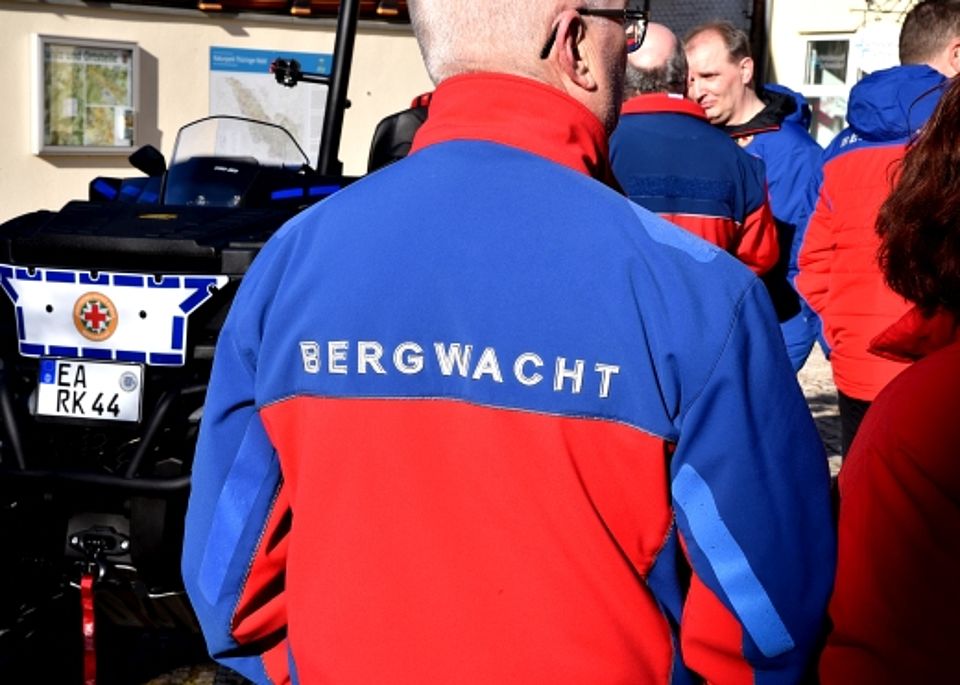 Rückenansicht eines Bergwacht-Mitglieds in rot-blauer Einsatzkleidung während einer Veranstaltung. Im Hintergrund steht das geländegängige ATV-Einsatzfahrzeug der Bergwacht.