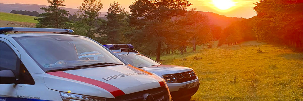 Zwei weiße Einsatzfahrzeuge der Bergwacht (eines mit dem Schriftzug "BERGWACHT" auf der Motorhaube) stehen bei Sonnenuntergang auf einer Lichtung oder einem Feld. Der Himmel und die hügelige, bewaldete Landschaft im Hintergrund sind in das warme, goldene Licht der untergehenden Sonne getaucht.
