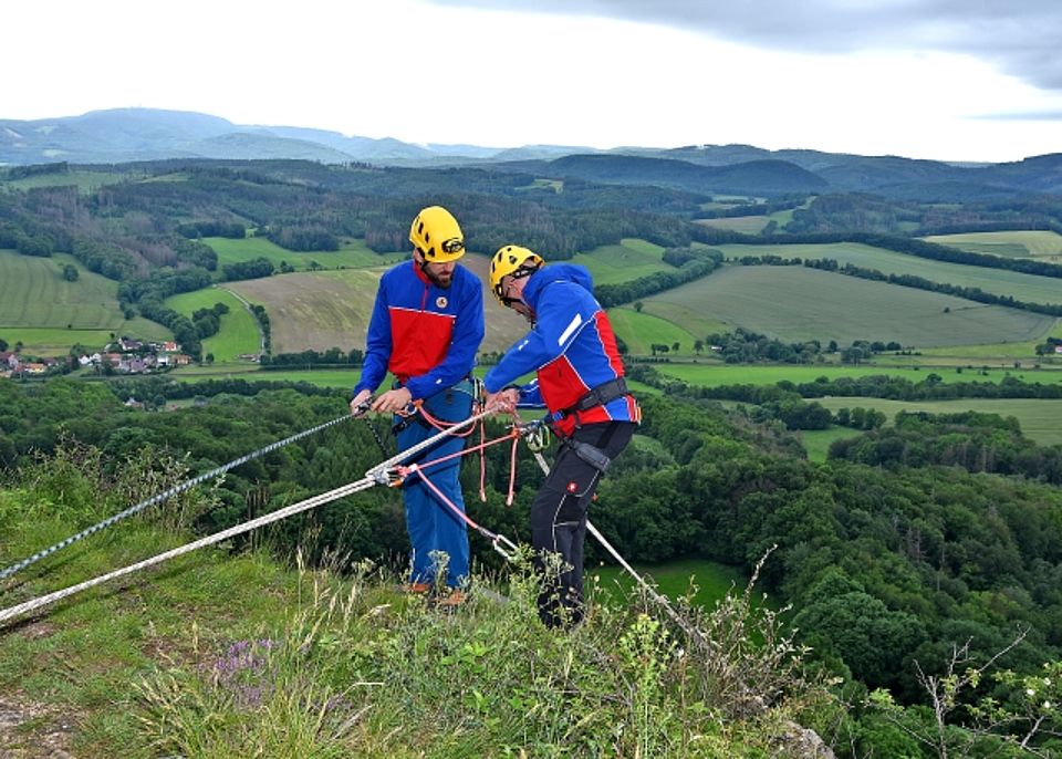 Zwei gesicherte Bergretter mit Helm seilen sich an einem Felsabhang ab. Im Hintergrund ist eine weite, grüne Landschaft zu sehen.