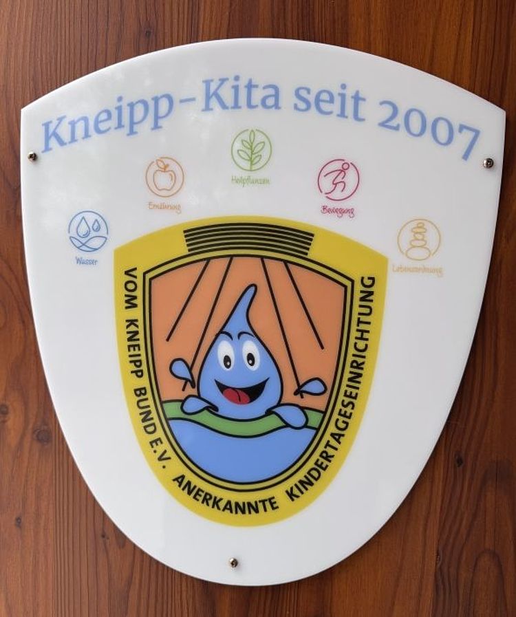 Das Bild zeigt ein weises Dreiecksschild. Oben steht der Text „Kneipp-Kita seit 2007“. Darunter sind fünf Symbole der Kneipp-Gesundheitslehre: Leben-Ordnung, Heilpflanzen, Bewegung, Ernährung und Wasser. In der Mitte befindet sich ein buntes Emblem mit einer lachenden blauen Wasserfigur vor stilisierten Sonnenstrahlen. Am unteren Rand steht „Vom Kneipp-Bund e. V. anerkannte Kindertageseinrichtung“.
