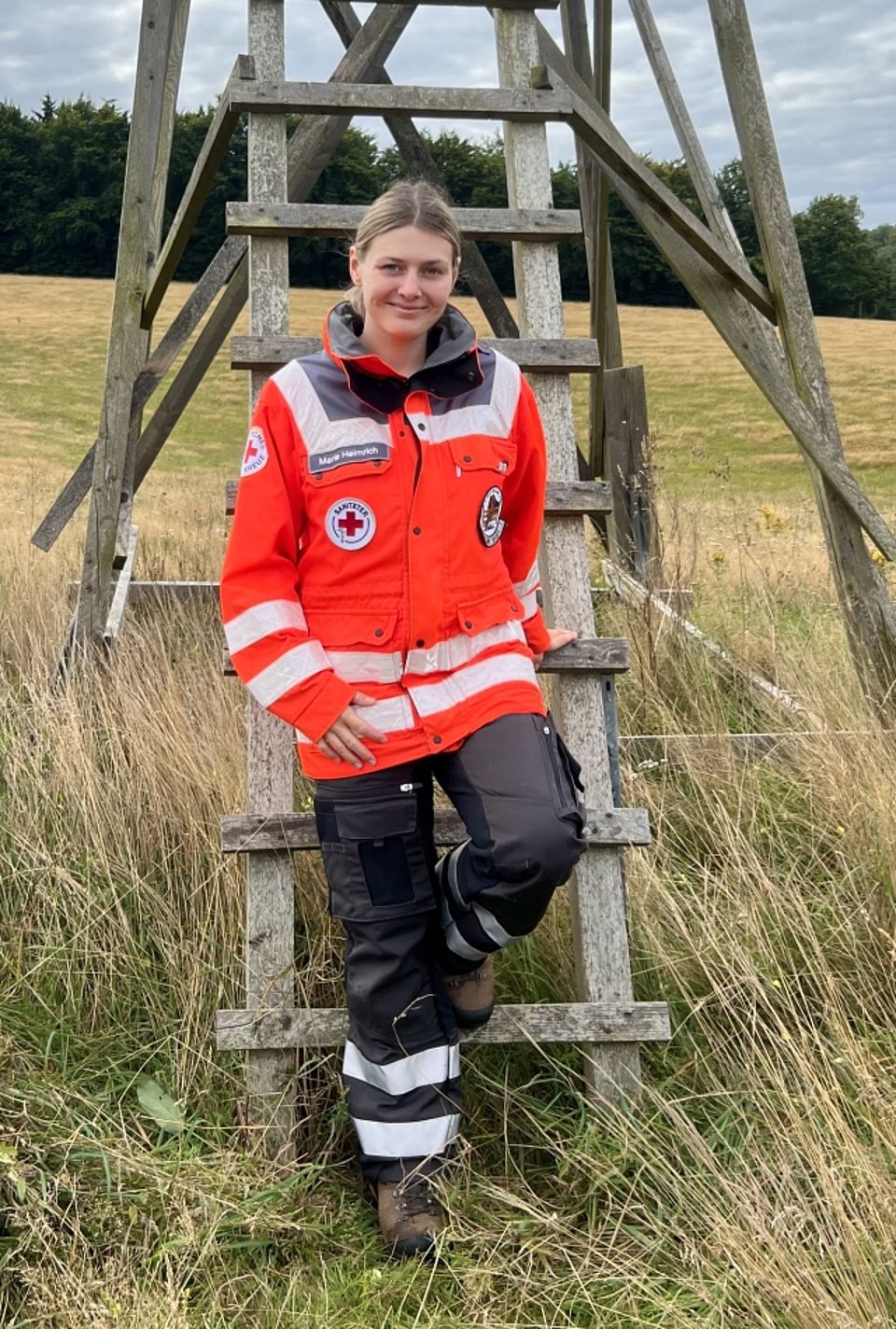 Marie Eine junge blonde Rettungskraft mit dem Namensschild "Marie -Christin Helmrich" steht gestützt auf einer rustikalen Holzleiter eines Hochsitzes in voller orange-grauer Einsatzkleidung des Deutschen Roten Kreuzes. Sie blickt lächelnd in die Kamera. Die Szene findet auf einer von hohem Gras umgebenen Wiese mit sanften Hügeln im Hintergrund unter einem bewölkten Himmel statt.