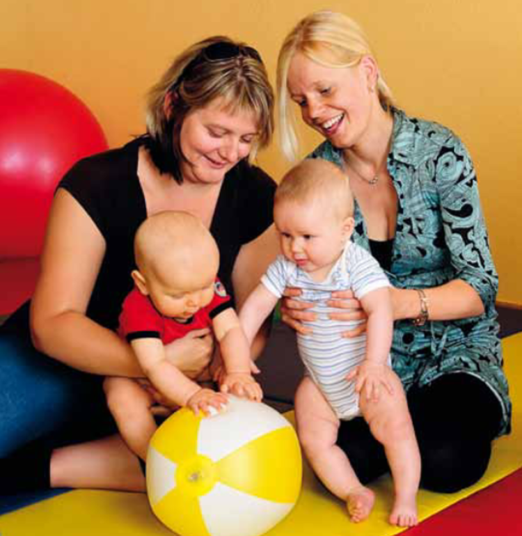 Zwei Frauen spielen mit ihren Kleinkindern und einem gelben Wasserball auf einer bunten Bodenmatte.