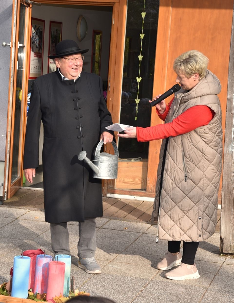 Ein als Sebastian Kneipp gekleideter Mann mit Gießkanne lacht neben einer Frau, die in ein Mikrofon spricht, vor dem Eingang des Regenbogenhauses.