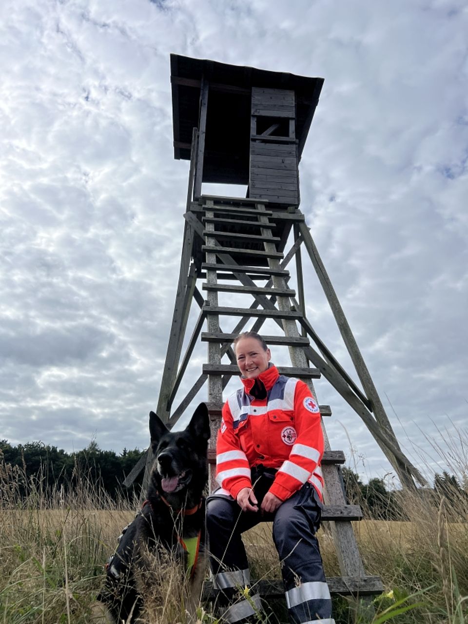 Annegreth mit Gizmo Eine lächelnde Frau (Rettungskraft) in orange-grauer Einsatzkleidung des Deutschen Roten Kreuzes sitzt auf einer Sprosse einer hohen, dunklen Holzleiter, die zu einem Hochsitz führt. Neben ihr sitzt ein großer, schwarzer Schäferhund Rettungshund mit abstehenden Ohren und geöffnetem Maul. Die Szene ist im Freien auf einer trockenen Wiese vor einem Waldrand und unter einem dramatisch bewölkten Himmel aufgenommen.