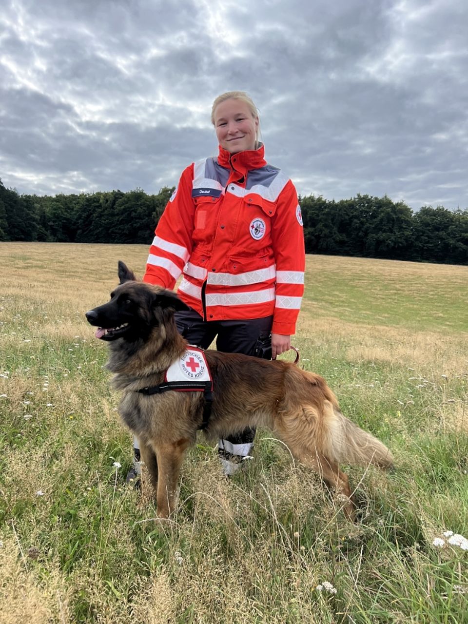 Isabel mit Ace Eine junge, blonde Frau (Rettungskraft mit dem Namensschild "Deubel") steht in orange-grauer Einsatzkleidung des Deutschen Roten Kreuzes auf einer Wiese. Sie lächelt in die Kamera. Neben ihr steht ein großer, langhaariger, braun-schwarz gefärbte Gelbbacke Rettungshund, der zur Seite blickt. Der Hund trägt ein weißes Geschirr mit dem Aufdruck "Rettungshundestaffel" und dem Roten Kreuz. Im Hintergrund ist ein Waldrand unter einem bewölkten Himmel zu sehen.