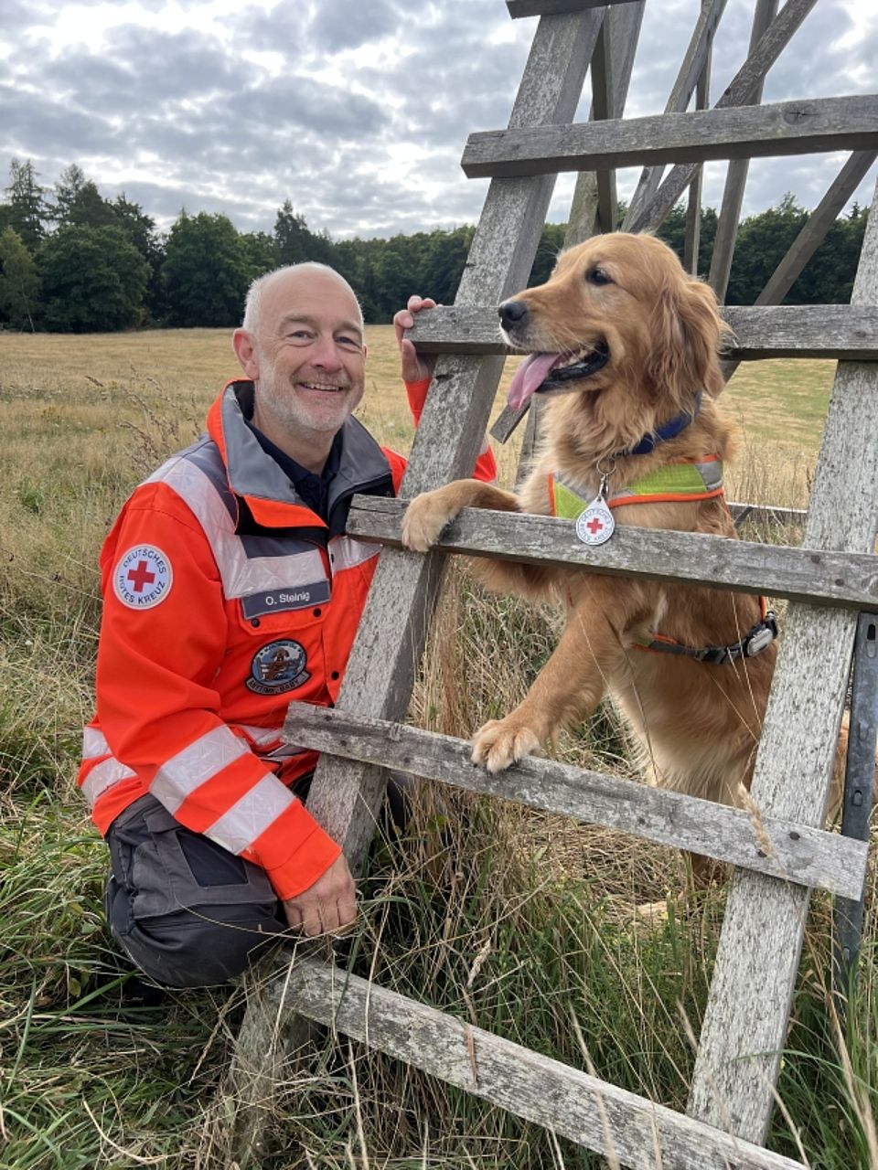 Olf mit Anouk Ein freundlich lächelnder, kurzhaariger Mann (eine Rettungskraft mit dem Namensschild "O. Steinig") kniet in orangefarbener Einsatzjacke des Deutschen Roten Kreuzes neben einem hellbraunen Golden Retriever Rettungshund. Der Hund steht mit den Vorderpfoten auf einer rustikalen Holzleiter oder einem Zaun und trägt eine reflektierende Weste mit einer Plakette des Roten Kreuzes. Die Szene spielt draußen auf einer Wiese mit Wald im Hintergrund.