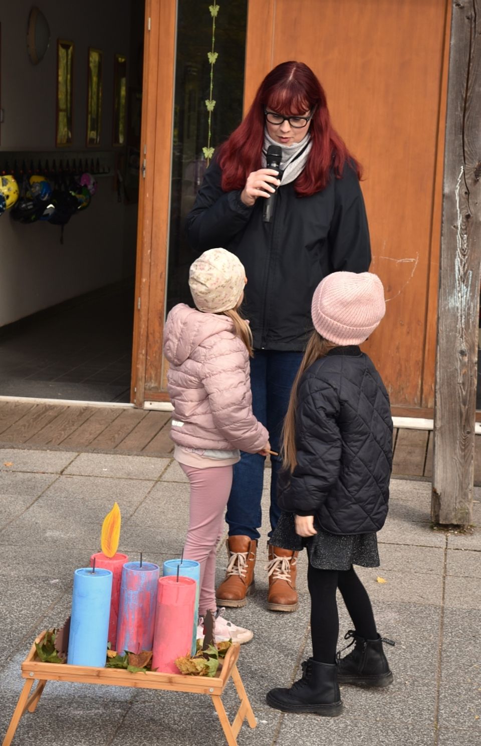 Eine Frau mit Mikrofon spricht im Beisein von zwei Kindern bei einer herbstlichen Veranstaltung vor dem Eingangsbereich; im Vordergrund stehen große, bunte Deko-Kerzen.
