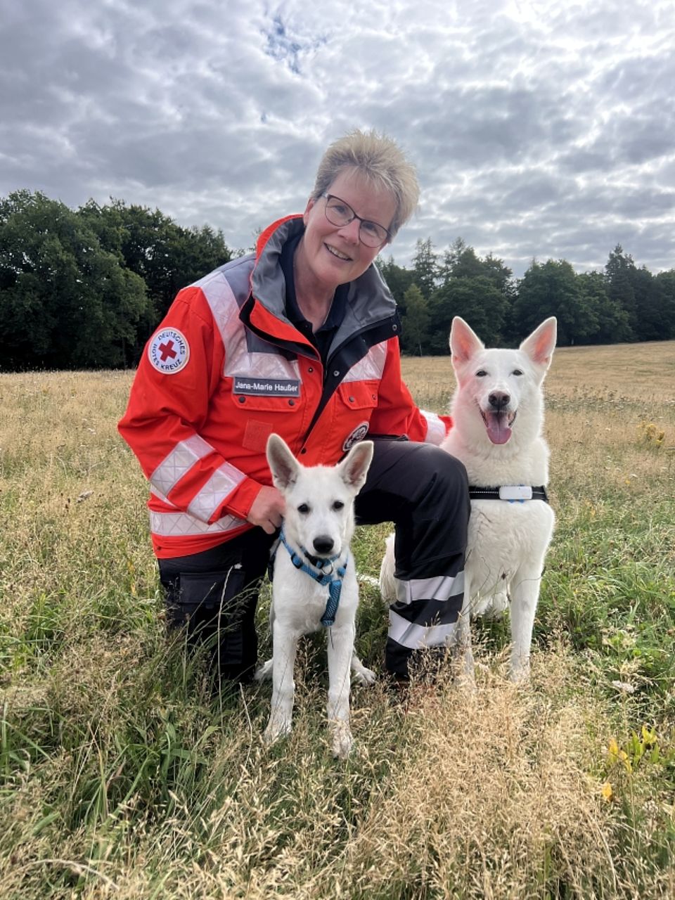 Jana mit Loki und Akira Eine lächelnde, kurzhaarige Frau (Rettungskraft mit dem Namensschild "Jana-Mary Haußer") in orange-grauer Einsatzkleidung des Deutschen Roten Kreuzes kniet zwischen zwei Weißen Schäferhunden Rettungshunden. Der Hund links ist ein Junghund mit blauer Leine, der direkt in die Kamera blickt. Der Hund rechts ist ein ausgewachsener Weißer Schäferhund, der mit offenem Maul ebenfalls in die Kamera blickt. Alle drei sind auf einer Wiese mit hohem Gras vor einem Waldrand unter bewölktem Himmel.