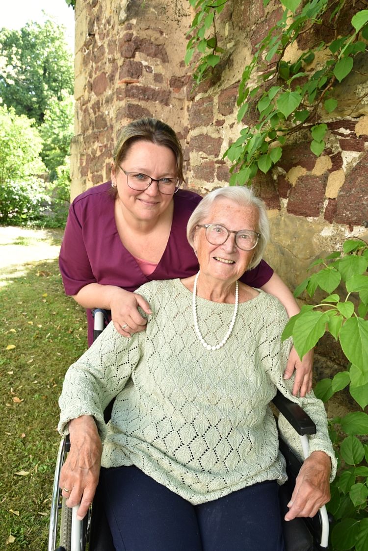 Eine ältere Frau sitzt in einem Rollstuhl und lächelt in die Kamera. Hinter ihr steht eine jüngere Pflegerin mit Brille, die ihr freundlich die Hände auf die Schultern legt. Beide befinden sich im Freien vor einer alten Steinmauer mit grünem Efeubewuchs.